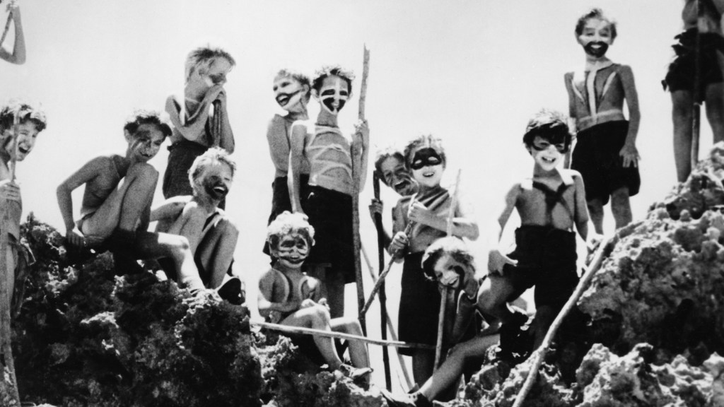 Black-and-white photograph of a group of young boys standing and crouching on rocky terrain, many with painted faces and holding spears, as if guarding something or about to go on a hunt.