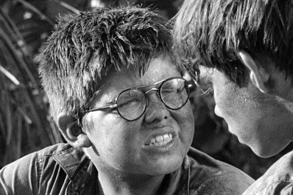 Black-and-white close-up of two boys facing each other outdoors in an island jungle. One wears round glasses and grimaces mid-sentence while the other listens closely as they share a confrontational discussion. 