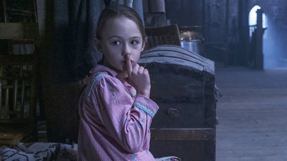A young girl in pink pajamas sits in a dimly lit attic beside an old wood trunk. She is holding one finger to her lips in a "shh" gesture and looking just off to the side of the camera.