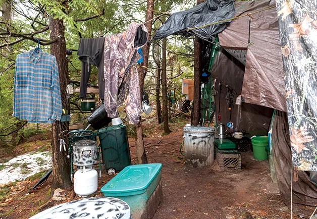 A makeshift campsite in a wooded forest. Tarps are strung between trees to form a shelter, with clothing hanging from lines. Buckets, plastic bins, a propane tank, pots, and other survival gear are scattered on the ground and tied to trees, showing Christopher Knight's living setup deep in the woods.