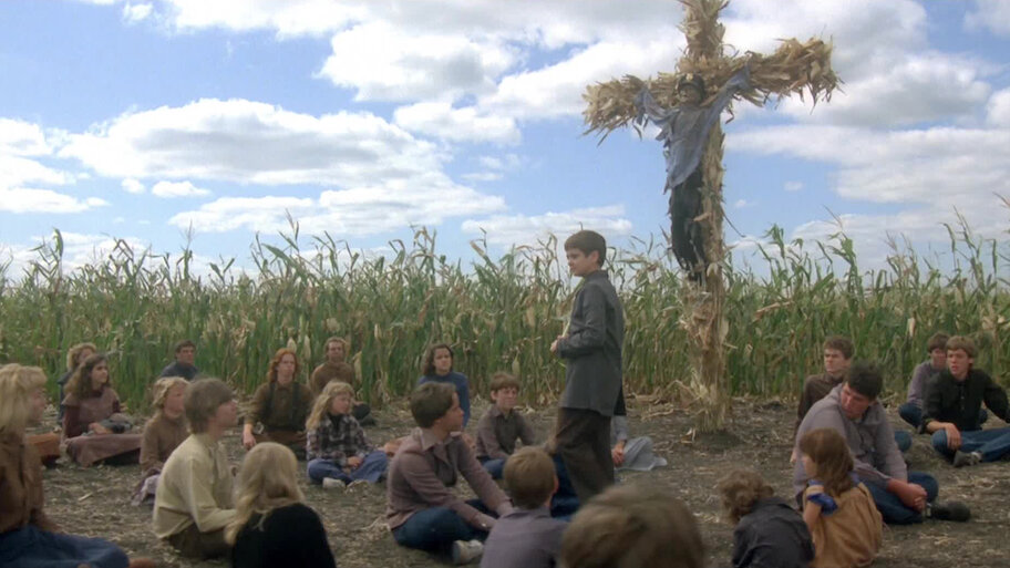 A scene from Children of the Corn. A group of children sit in a circle at the edge of a cornfield. A boy stands at the center. Behind him is a police officers skeleton perched as a scarecrow attached to a tall wooden cross made of dried corn stalks.