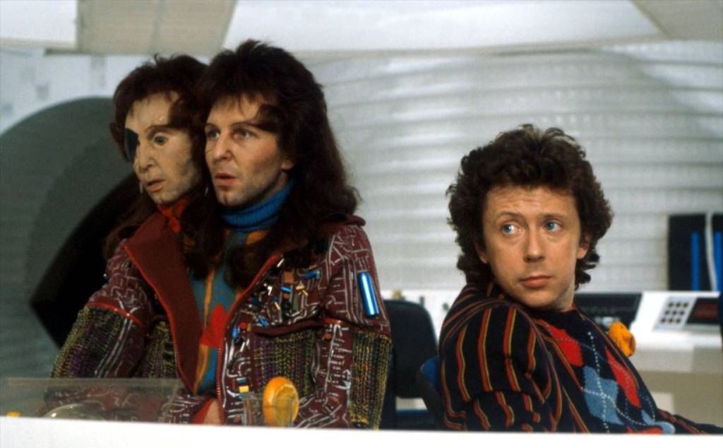 A still from the 1981 BBC miniseries showing Zaphod Beeblebrox with his prosthetic second head visible over his left shoulder, seated next to Arthur Dent in the cockpit of their spaceship. Both are wearing colorful, eclectic 1980s sci-fi costuming.