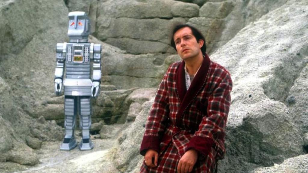 Arthur Dent, wearing a plaid bathrobe, sits on a rock beside Marvin the Paranoid Android — a boxy, silver robot standing to his left. Both look off into the distance.