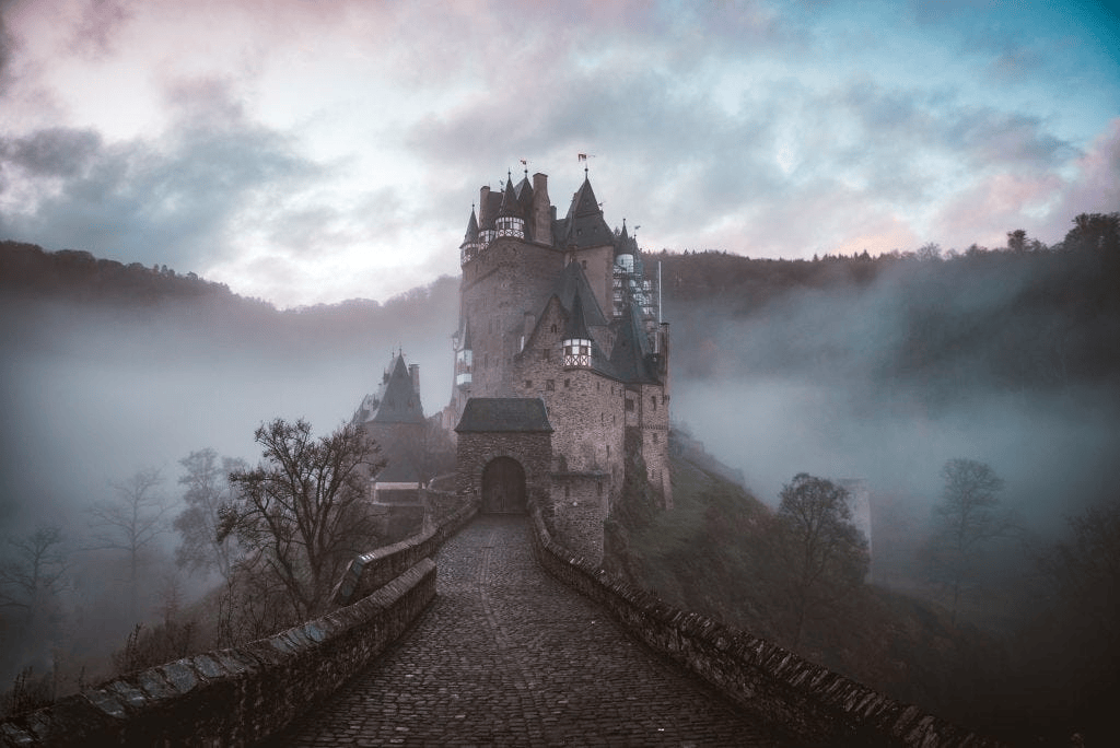 A stone bridge leads to the entrance of Eltz Castle, a medieval fortress rising out of a dense, low-lying fog under a dramatic, cloudy sky.