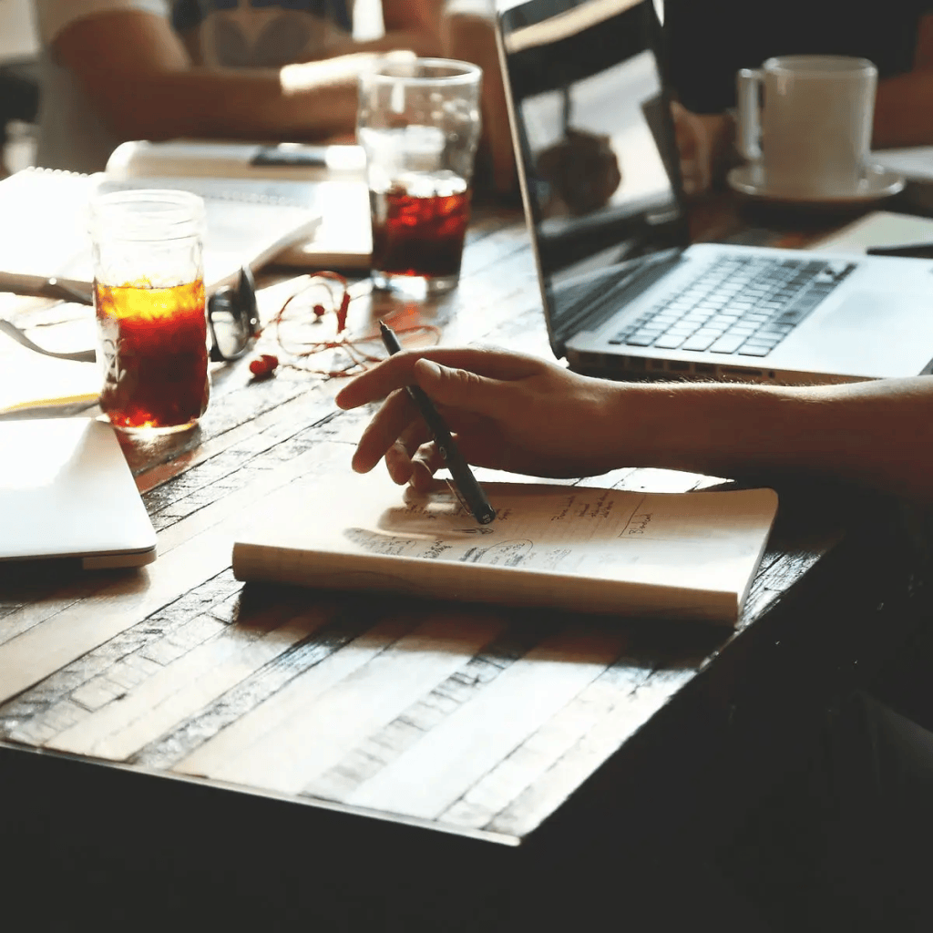 A close-up shot of a person’s hand writing with a pen in a notebook on a dark, rustic wooden table. An open laptop, a glass of iced coffee, eyeglasses, and a white coffee mug are scattered across the desk, creating a busy collaborative work environment.